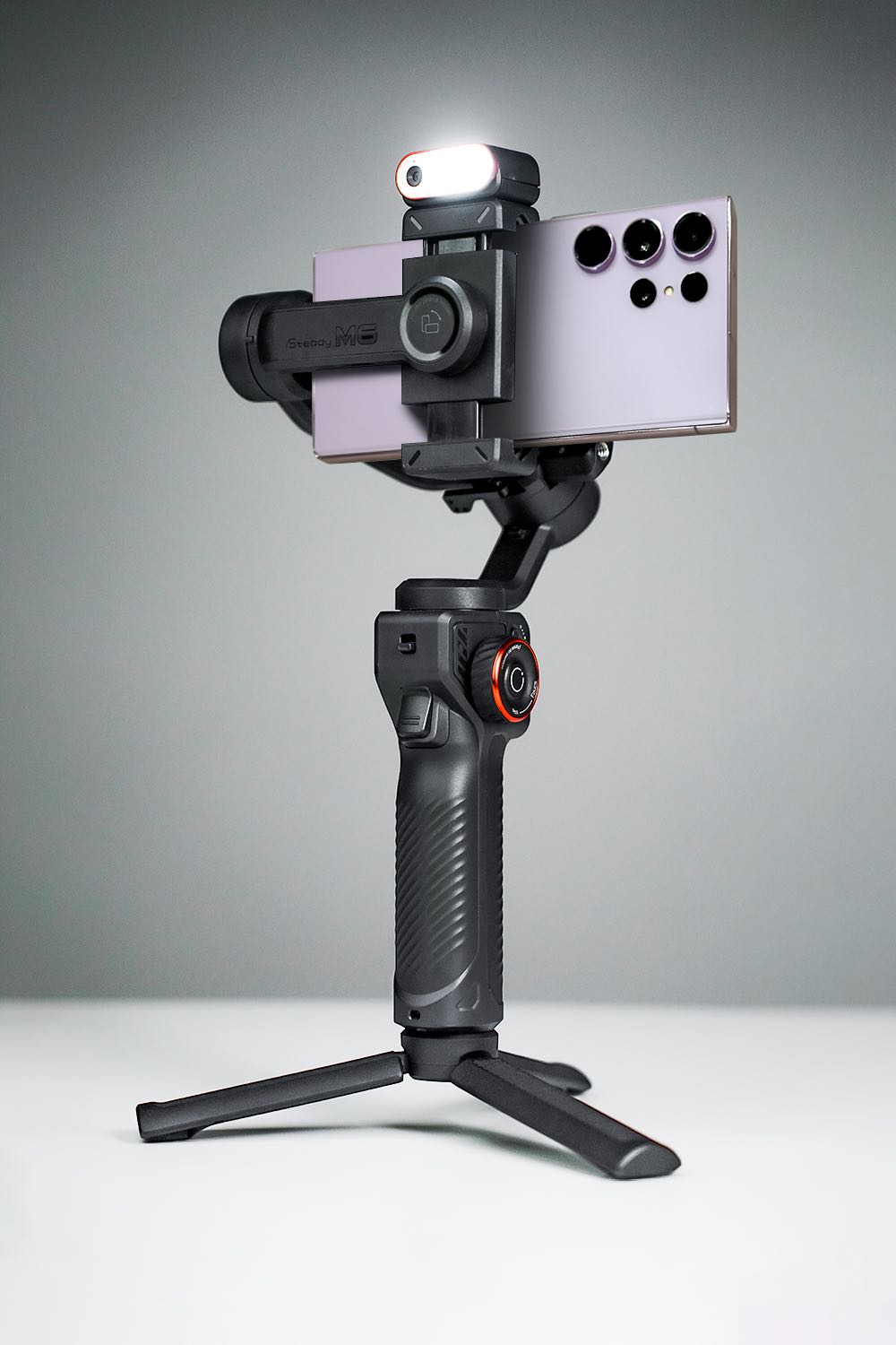 Best gimbal for Samsung S23 and S24 Ultra: A black handheld gimbal with a mounted S24 Ultra smartphone and an LED light on top, set against a neutral gray background.