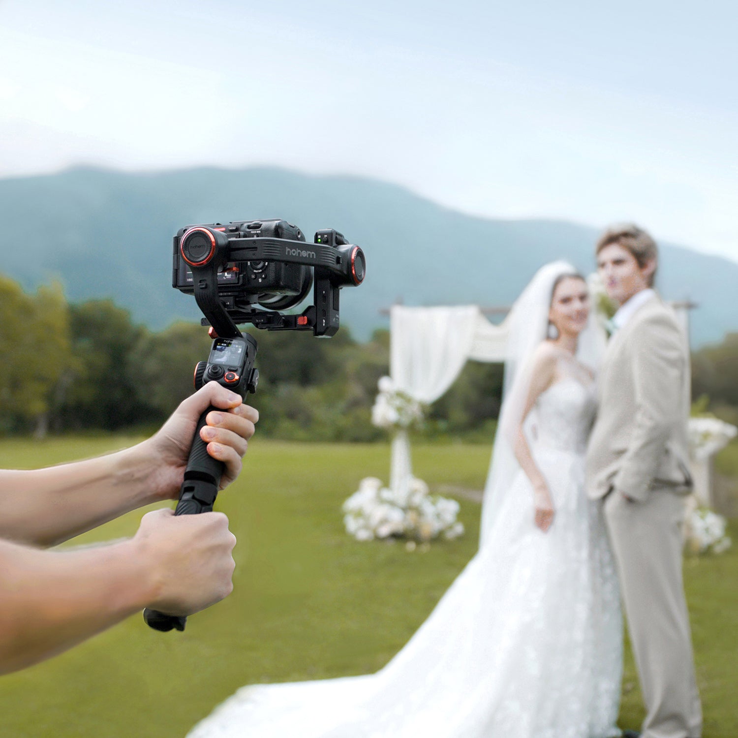 Person filming a bride and groom with a camera stabilizer outdoors.