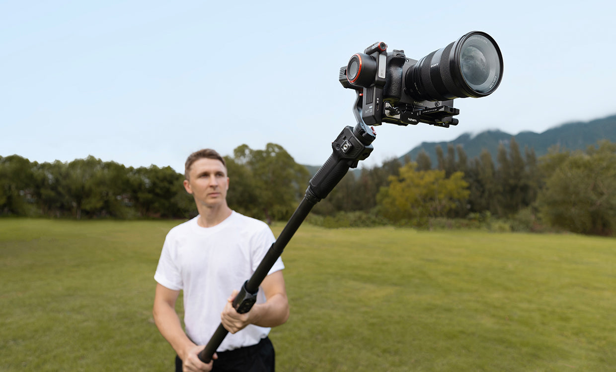 Man holding a camera on a stabilizer in an outdoor setting with trees and mountains.